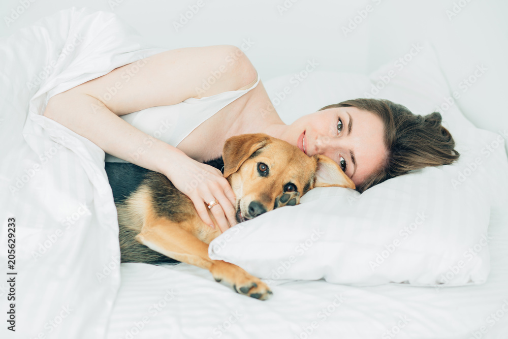 Beautiful excited young woman and her cute cur dog are fool around, looking at camera while lying covered with a blanket in bed. White background. Pet lover concept. Beautiful excited young woman and her cute cur dog are fool around, looking at camera while lying covered with a blanket in bed. White background. Pet lover concept.