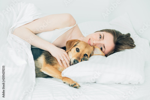 Beautiful excited young woman and her cute cur dog are fool around, looking at camera while lying covered with a blanket in bed. White background. Pet lover concept.