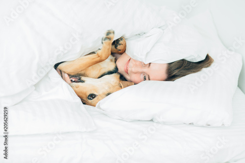 Beautiful excited young woman and her cute cur dog are fool around, looking at camera while lying covered with a blanket in bed. White background. Pet lover concept.