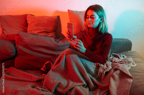 Portrait of young lady sittig with plaid and using cellphone on sofa while spending time at home