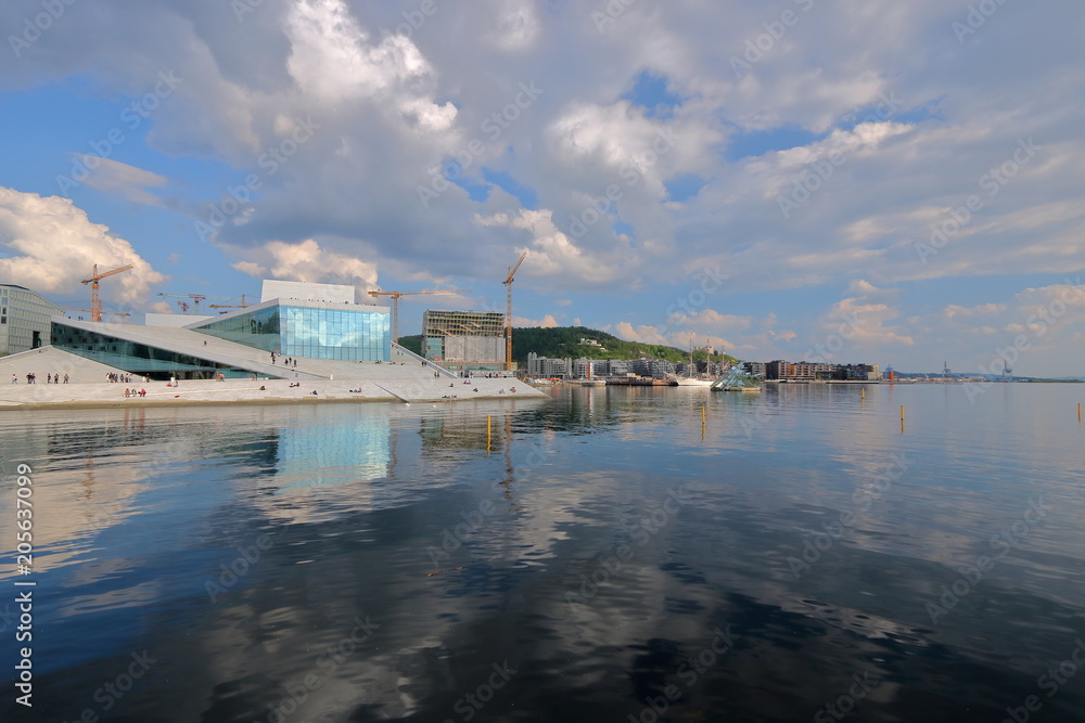 Widok budynku opery w Oslo, Norwegia, z przeciwnego brzegu zatoki morza, promenady z nowymi budynkami na brzegu, słonecznie, malownicze chmury na niebie