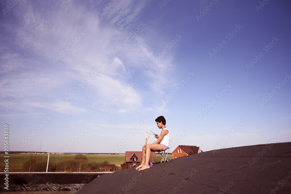 Boy sitting on the roof of the house and shoots from slingshot Boy sitting on the roof of the house and shoots from slingshot