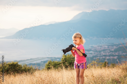 Little kid is playing with camera and taking photo of sea and mountains as master. Blonde girl is traveling photographer.