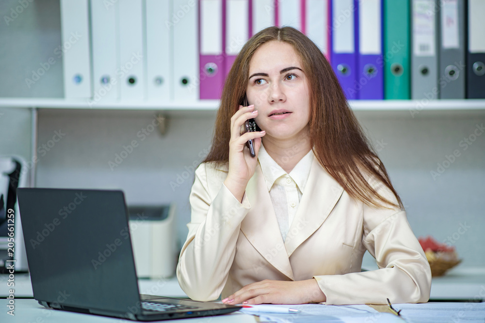 Portrait of a beautiful brunette manager girl working in the office with a laptop and talking on a smartphone. Portrait of a beautiful brunette manager girl working in the office with a laptop and talking on a smartphone.