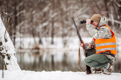 Male hunter in camouflage, armed with a rifle, standing in a snowy winter forest with duck prey