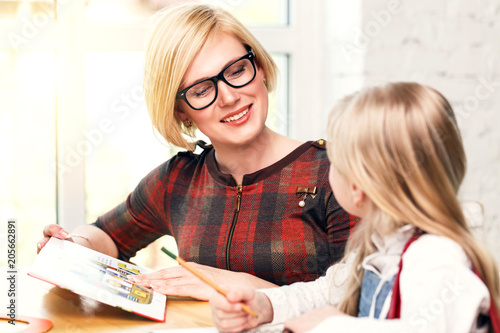 Pretty smiling blonde English teacher wears eyeglasses studying with kid girl, language learning with children in white light classroom