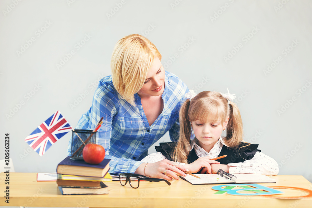 Pretty adult female teacher helps kid girl to learn English language, school table with books, flag and letters in light classroom Pretty adult female teacher helps kid girl to learn English language, school table with books, flag and letters in light classroom