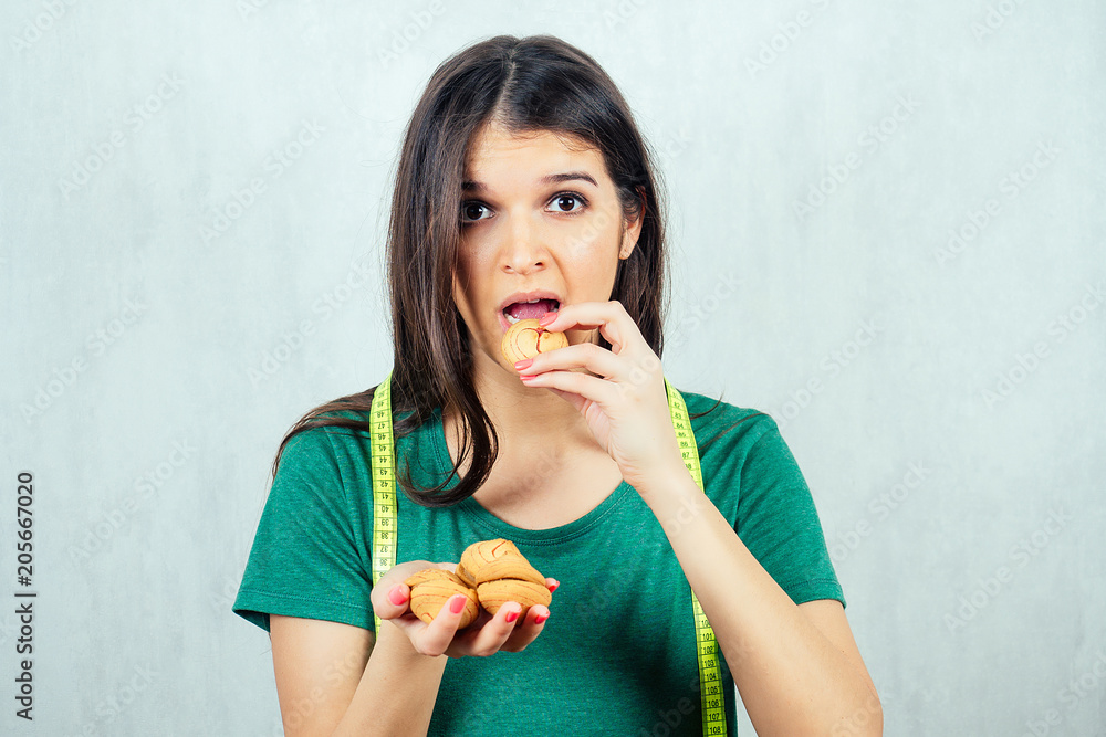 portrait of a hungry, young and beautiful woman holding a cookie in a T-shirt and measuring tape. concept of diet and rejection of unhealthy harmful food