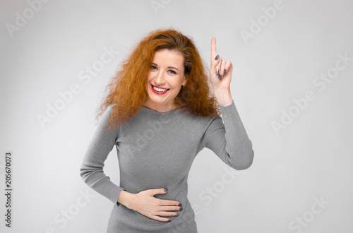 Charming young girl with red hair. A young girl in a gray sweater. Young girl on a gray background. A young girl holds her hand behind her belly. Young girl showing thumb up photo