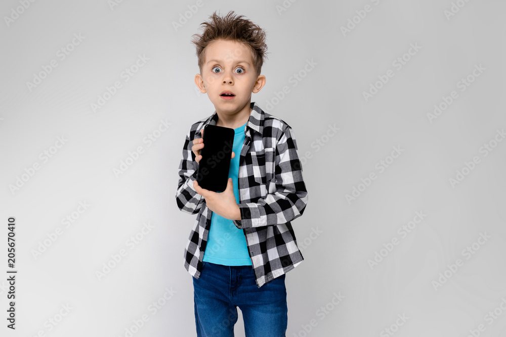 A handsome boy in a plaid shirt, blue shirt and jeans stands on a gray background. The boy is holding a phone A handsome boy in a plaid shirt, blue shirt and jeans stands on a gray background. The boy is holding a phone