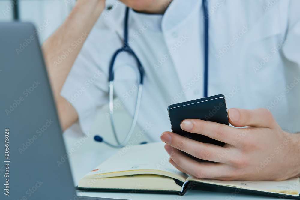 Close up male doctor or medical student using mobile smart phone working on laptop computer in hospital. Close up male doctor or medical student using mobile smart phone working on laptop computer in hospital.