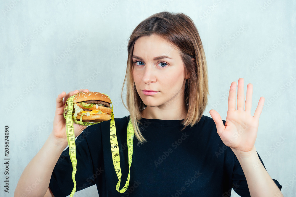 young and attractive blond woman in black T-shirt and measuring tape is holding a high-calorie burger and shows a hand gesture stop. concept of refusing harmful fast food and diet young and attractive blond woman in black T-shirt and measuring tape is holding a high-calorie burger and shows a hand gesture stop. concept of refusing harmful fast food and diet