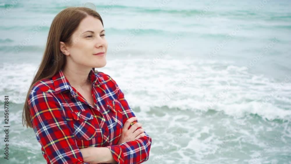 Young woman looking at big sea waves on beach. Female traveler is cold during vacation on beach at stormy weather while off-season. Breeze from ocean blowing her hair in slow motion Young woman looking at big sea waves on beach. Female traveler is cold during vacation on beach at stormy weather while off-season. Breeze from ocean blowing her hair in slow motion