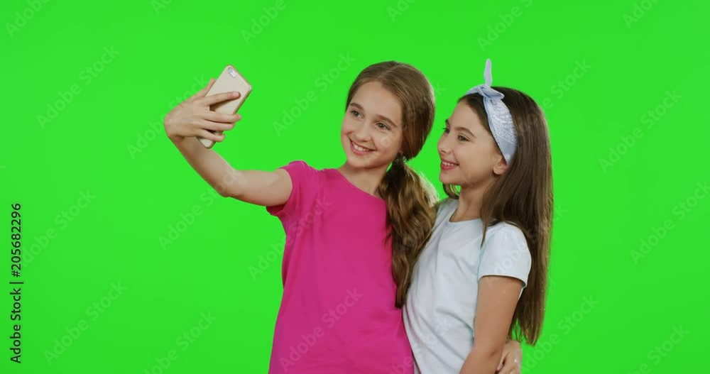 Caucasian friendly teens girls making selfies on the smartphone and posing while standing on the green screen background. Chroma key Caucasian friendly teens girls making selfies on the smartphone and posing while standing on the green screen background. Chroma key