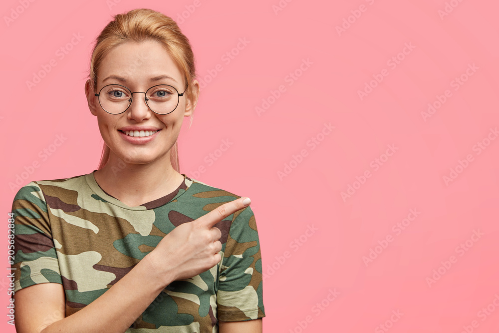 Cheerful blonde female student uses copy space for advertising, points right and smiles broadly, demonstrates her project or creative work, wears casual clothes, isolated over pink background Cheerful blonde female student uses copy space for advertising, points right and smiles broadly, demonstrates her project or creative work, wears casual clothes, isolated over pink background