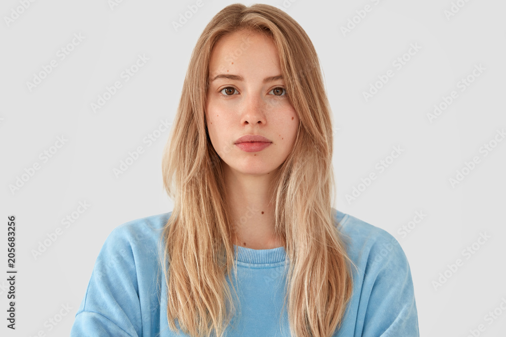 Headshot of tender beautiful young female model with fair hair, wears blue top, has healthy pure skin, looks seriously at camera, poses against white concrete wall. Cute teenager feels confident Headshot of tender beautiful young female model with fair hair, wears blue top, has healthy pure skin, looks seriously at camera, poses against white concrete wall. Cute teenager feels confident