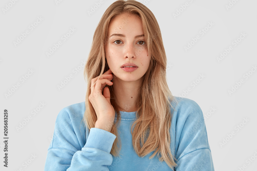 Portrait of beautiful European femae with tender look, has pleasant appearance and healthy skin, poses indoor against white background, wears casual long sleeved light blue sweater. People, lifestyle Portrait of beautiful European femae with tender look, has pleasant appearance and healthy skin, poses indoor against white background, wears casual long sleeved light blue sweater. People, lifestyle
