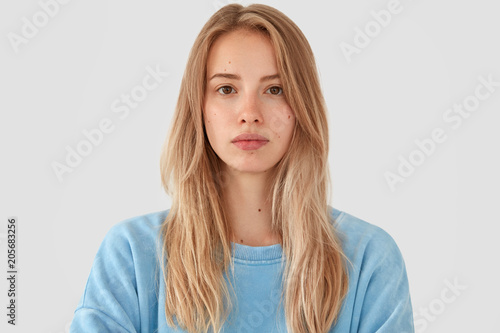 Headshot of tender beautiful young female model with fair hair, wears blue top, has healthy pure skin, looks seriously at camera, poses against white concrete wall. Cute teenager feels confident