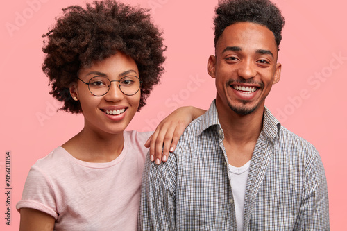 Photo of friendly African American male and female stand close to each other, have broad smiles on faces, have date together, pose against pink background. Beautiful dark skinned woman and her friend