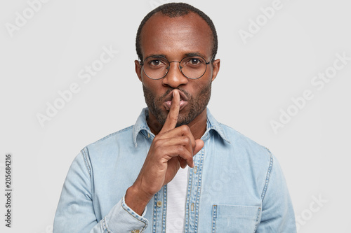 Keep this in secret! Serious dark skinned man keeps voice down, shows silence sign with index finger over mouth, asks to be quiet, wears round spectacles, isolated on white concrete background