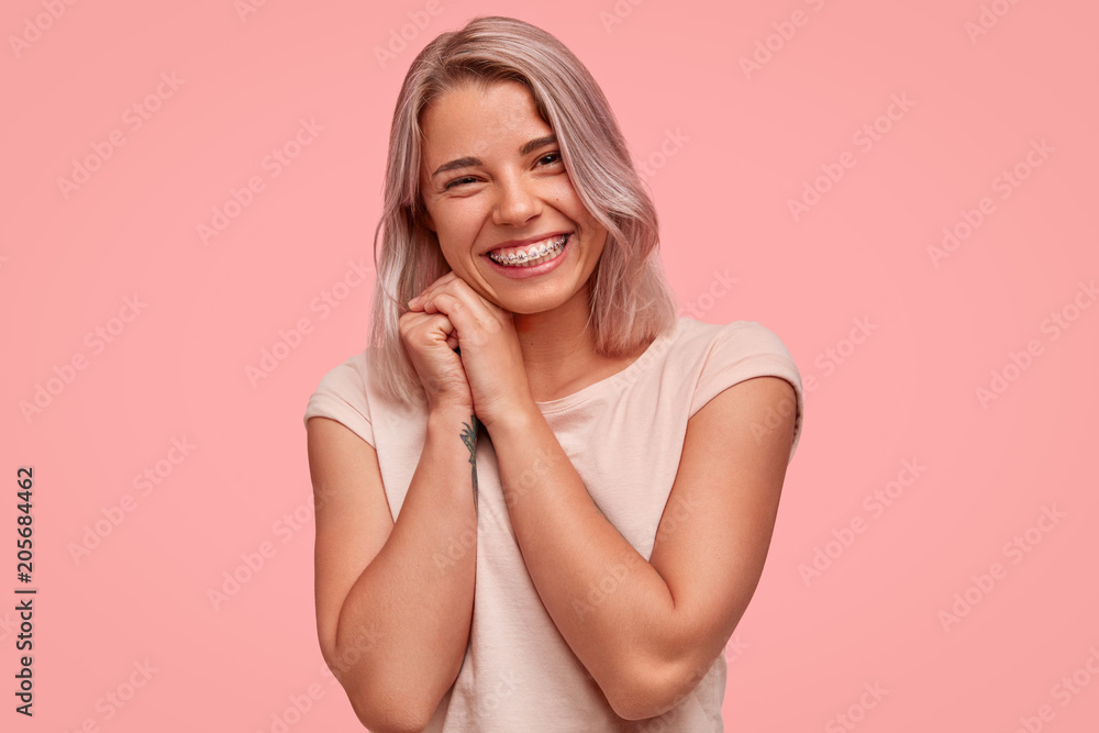 Carefree beautiful female with braces on teeth, smiles broadly at camera, being in high spirit while hangs out with friends, has joyful expression, stands against pink background. Optimistic girl Carefree beautiful female with braces on teeth, smiles broadly at camera, being in high spirit while hangs out with friends, has joyful expression, stands against pink background. Optimistic girl