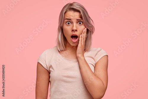 Waist up portrait of stunned woman drops jaw in surprisement, being shocked with curious revelation, hears bad news, wears light casual t shirt in one tone with background. Monochrome concept