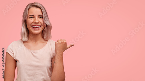This is perfect place for entertaining. Positive good looking female model with bobbed hairstyle, indicates with thumb at blank copy space, raises hand, blank pink wall for your advertisement photo