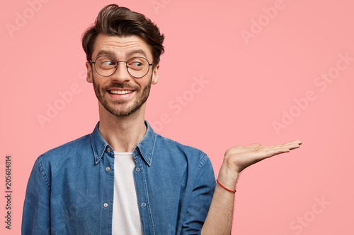 Cheerful attractive male with rendy hairstyle, being in good mood, shows with palm city banner for friends, feels proud to be involved in advertisement, poses against pink background, blank space photo