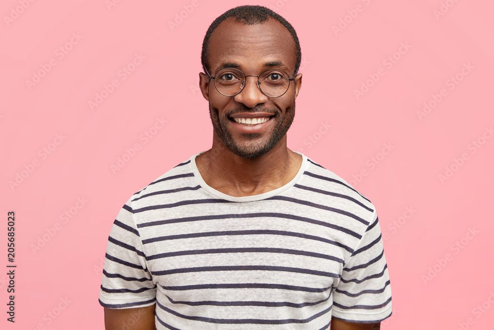 Handsome delighted guy with dark skin, pleasant smile, wears casual striped t shirt, poses against pink background, expresses positive emotions and feelings. People, facial expressions concept Handsome delighted guy with dark skin, pleasant smile, wears casual striped t shirt, poses against pink background, expresses positive emotions and feelings. People, facial expressions concept