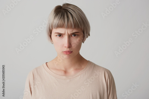 indoor shot of young female looks serious with confident expression, feels angry and mad. isolated over white studio wall