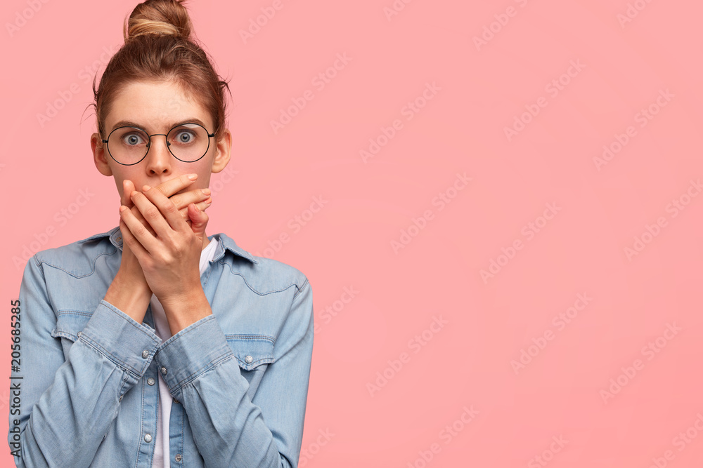 Portrait of attractive female being shocked with prices during shopping, covers mouth with palms, expresses great surprisement, wears denim jacket, stands against pink background, blank space