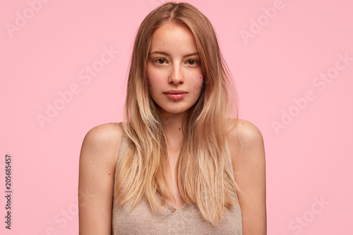 Beautiful young dark eyed female model with pleasant apperance, long straight hair, poses at camera with serious expression, reads something attentively on billboard, isolated on pink background