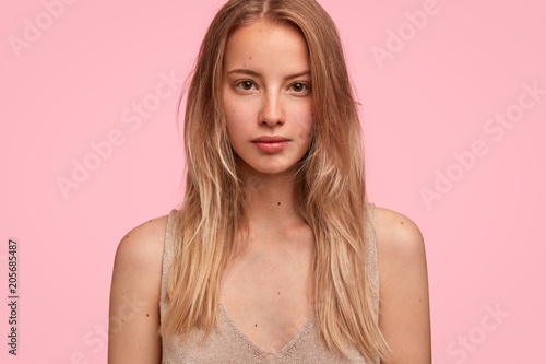 Cropped shot of lovely young woman being focused at camera, has pleasant appealing look, long hair, tries to hear necessary information, poses against pink background. People, beauty concept