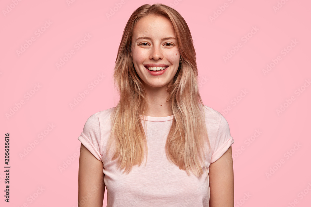 Studio shot of pleasant looking young female with charming smile, being delighted to see something wonderful or meet with close friend, dressed in casual outfit, stands against pink background Studio shot of pleasant looking young female with charming smile, being delighted to see something wonderful or meet with close friend, dressed in casual outfit, stands against pink background