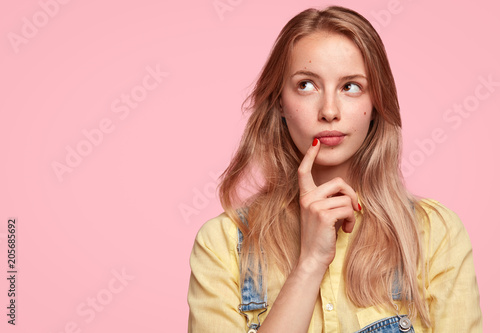 Thoughtful female youngster keeps fore finger near lips, being deep in thoughts, thinks about coming holidays, isolated on pink background with blank copy space for your advertisement or text