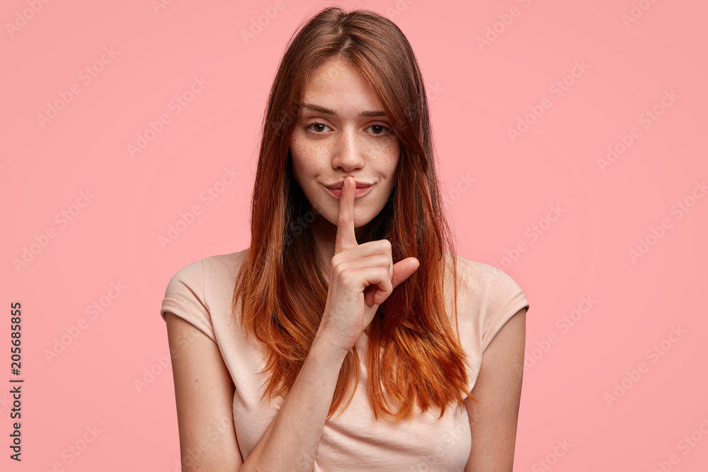 Silence concept. Attractive young female model with freckled healthy skin, keeps index finger on lips, asks to be mute as someone is sleeping, has serious expression, poses against pink background Silence concept. Attractive young female model with freckled healthy skin, keeps index finger on lips, asks to be mute as someone is sleeping, has serious expression, poses against pink background