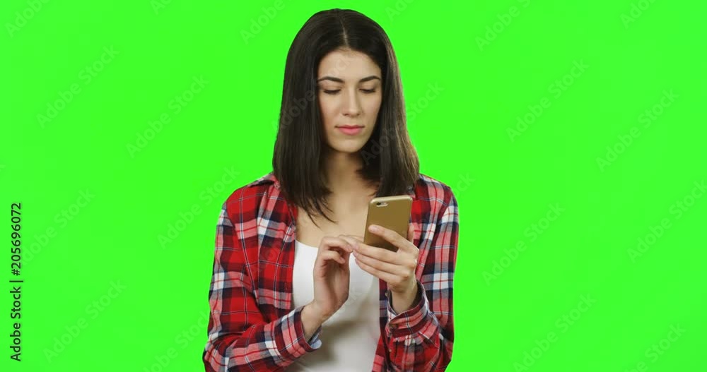 Portrait shot of the brunette young good looking woman in the plaid red shirt scrolling and taping on the smartphone in front of the camera on the green background. Chroma key Portrait shot of the brunette young good looking woman in the plaid red shirt scrolling and taping on the smartphone in front of the camera on the green background. Chroma key