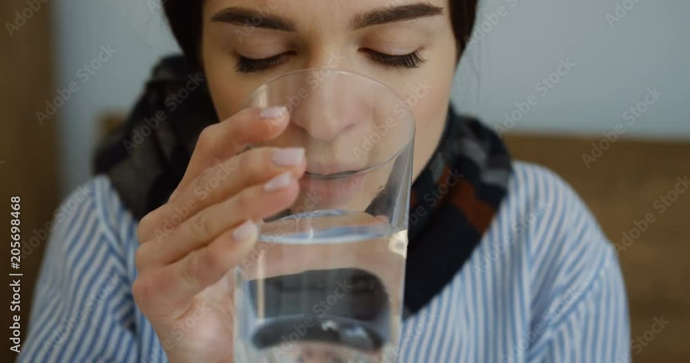 Close up of the unhealthy young Caucasian woman in a scarf and pajama taking a pill and drinking water as having a flu. Inside Close up of the unhealthy young Caucasian woman in a scarf and pajama taking a pill and drinking water as having a flu. Inside