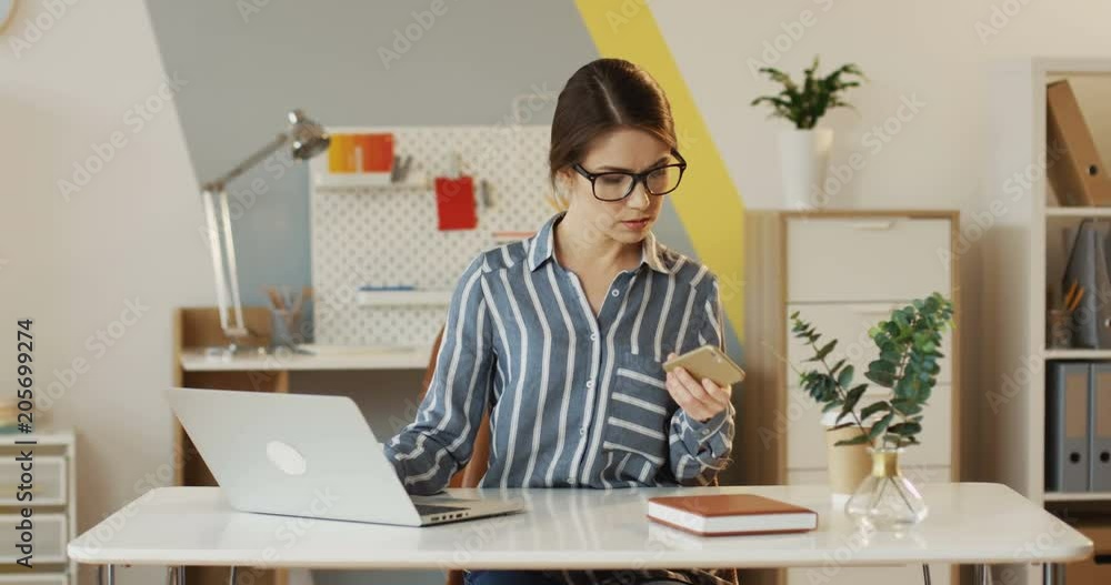 Beautiful Caucasian woman in the striped blouse and glasses sitting at the table with a laptop computer and talking on the phone while working in the office. Indoor. Beautiful Caucasian woman in the striped blouse and glasses sitting at the table with a laptop computer and talking on the phone while working in the office. Indoor.
