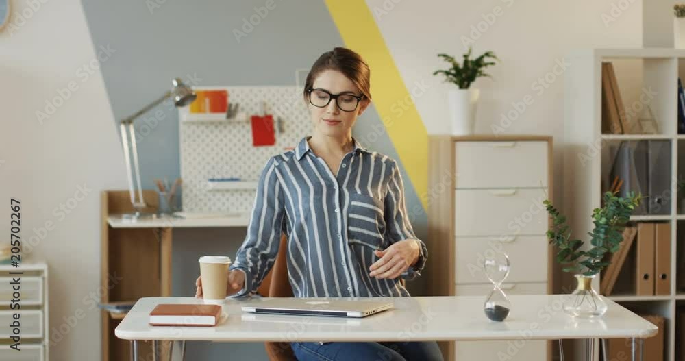 Pretty young woman in the glasses coming to her workplace with coffee, sitting at the table, opening her laptop, starting to work and smiling to the camera. Inside Pretty young woman in the glasses coming to her workplace with coffee, sitting at the table, opening her laptop, starting to work and smiling to the camera. Inside