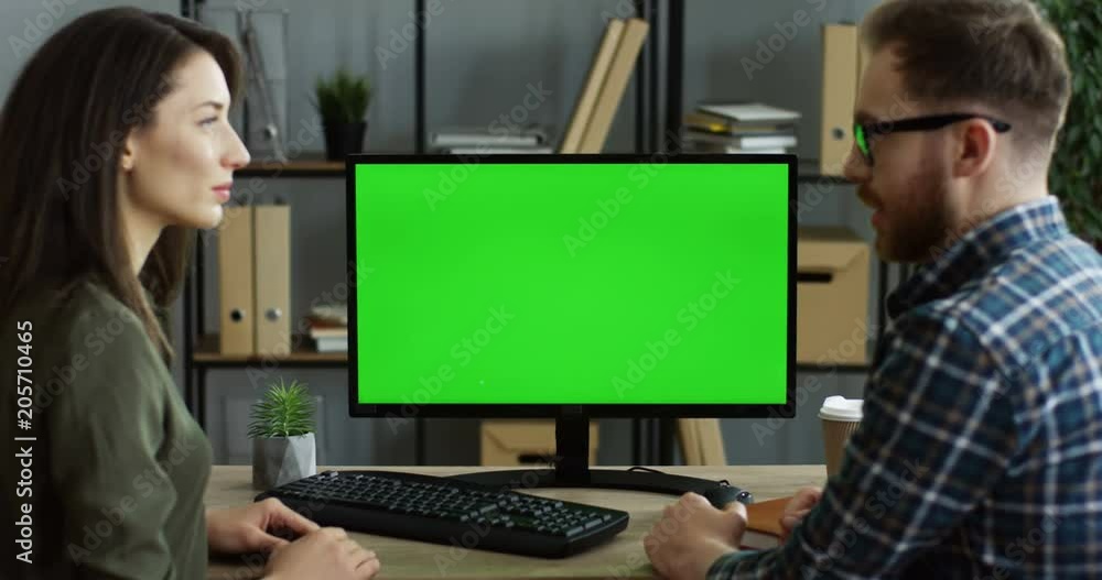 View from the back on the man and woman sitting at the office table half turning to the camera and looking at each other and at the computer green screen in focus. Indoor View from the back on the man and woman sitting at the office table half turning to the camera and looking at each other and at the computer green screen in focus. Indoor