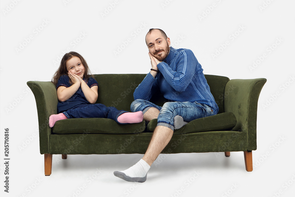 Bedtime. Picture of attractive positive young man in jeans and sweater sitting on sofa in living room, trying to send his daughter to bed, placing hands under cheek and smiling joyfully at camera