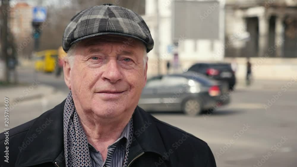 Portrait of a cheerful senior man in a chechered cap smiling and standing in a street with traffic in the background in slow motion Portrait of a cheerful senior man in a chechered cap smiling and standing in a street with traffic in the background in slow motion