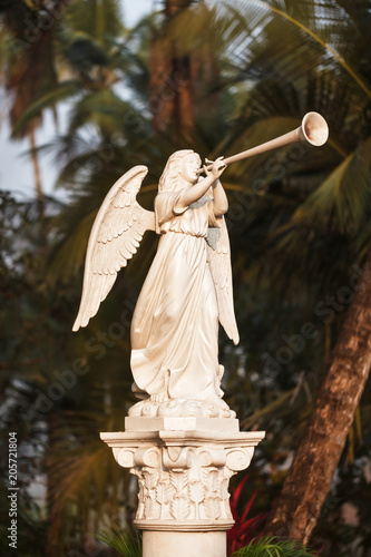The angel in white with big wings blows the trumpet in dawn beams - a sculpture near Catholic church in front of the background of tropical trees and palms.
