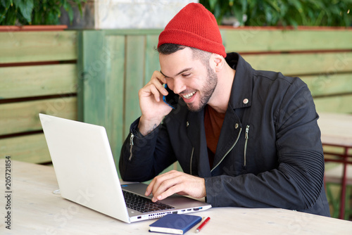 funny young hipster man laughs merrily while texting on a laptop and talking on the phone. The concept of lifestyle mobility