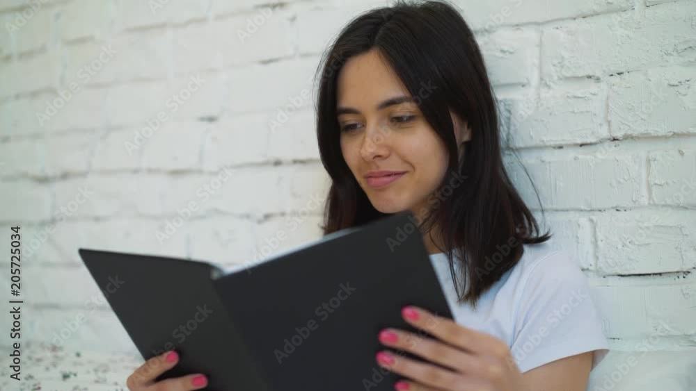 Beautiful brunette girl sitting on the bed holding a book to read. A young woman opens an interesting book. Beautiful brunette girl sitting on the bed holding a book to read. A young woman opens an interesting book.
