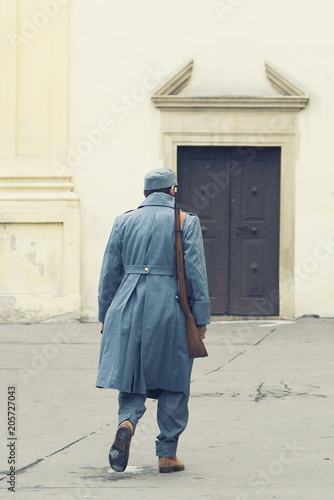 Soldier of Austro-Hungarian Army with a rifle on his shoulder walking into a closed roman Catholic church s door. Winter grey uniform against yelow facade © vlad