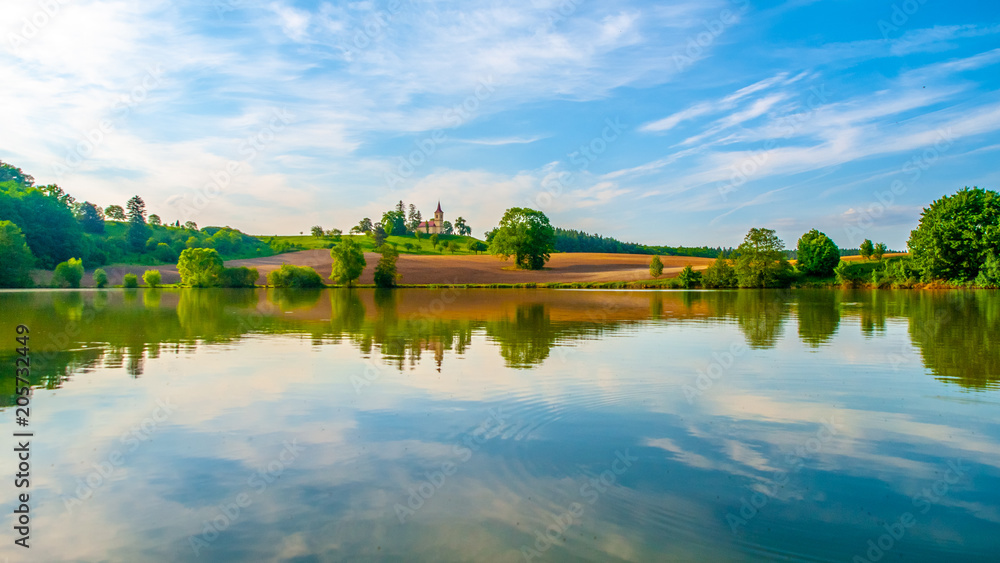 Romantic landscape with small church on the hill reflected in the pond. Sunny summer day with blue sky and white clouds. St. Peter and Pauls church at Bysicky near Lazne Belohrad, Czech Republic.