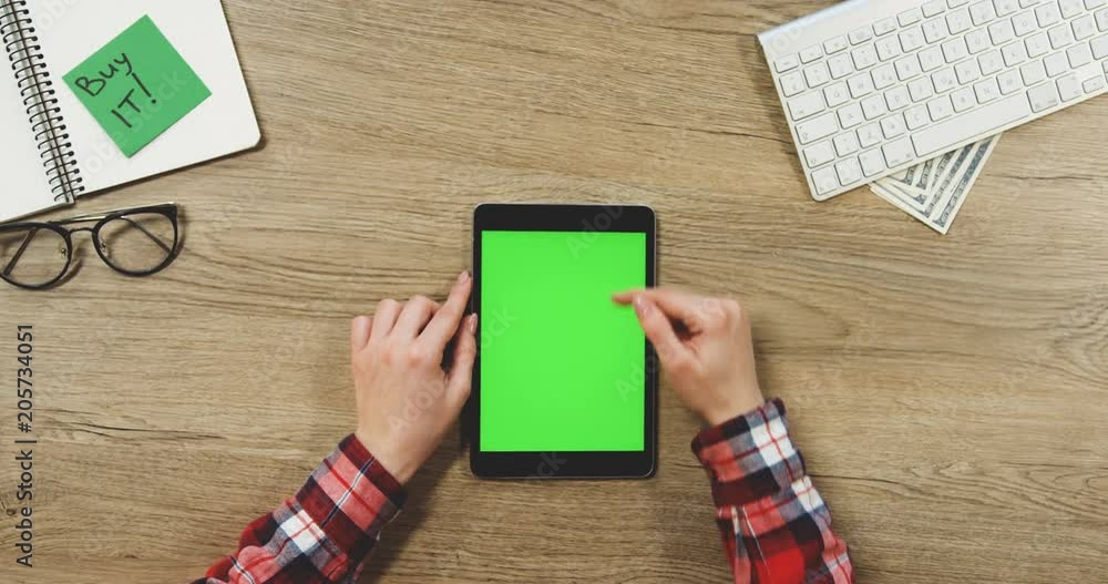View from above on the female hands using the black vertical tablet device with a green screen on the wooden desk. Buy it card, glasses and keyboard. Chroma key. Close up View from above on the female hands using the black vertical tablet device with a green screen on the wooden desk. Buy it card, glasses and keyboard. Chroma key. Close up