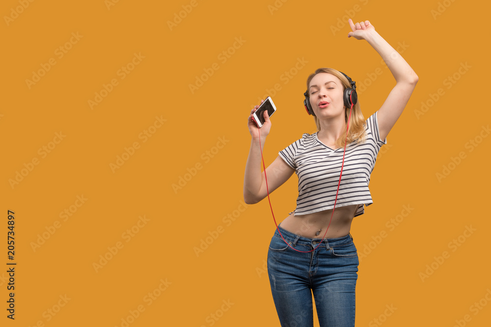 A cheerful hipster girl listening to music on headphones with a phone on a yellow background
